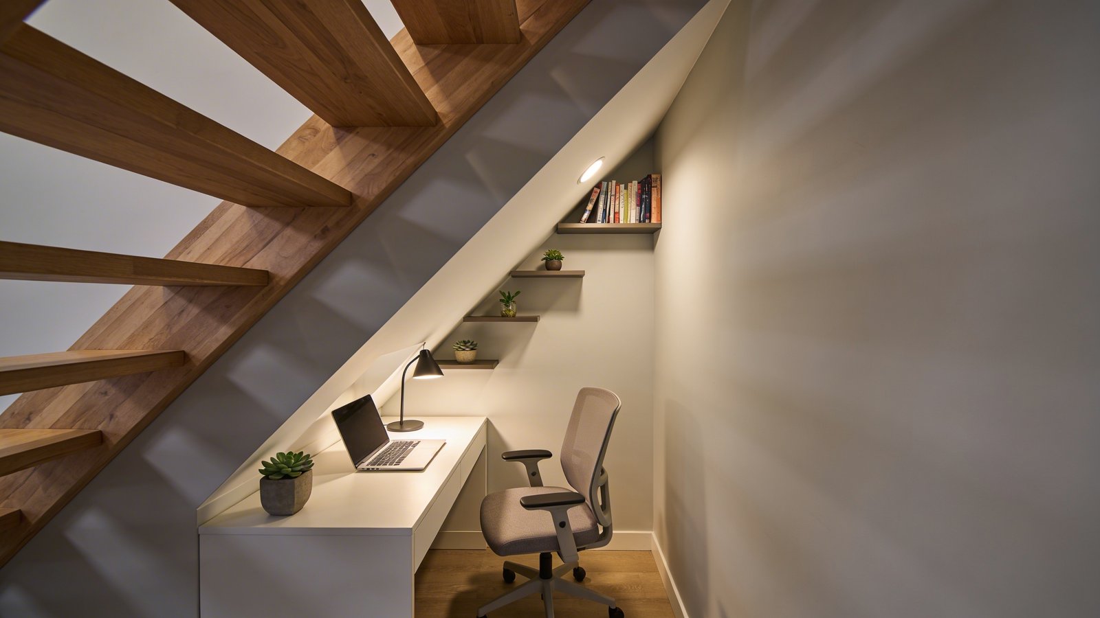 A cozy home office cleverly built into the triangular space beneath a wooden staircase, featuring a modern white desk with a laptop and desk lamp tucked perfectly into the slanted area, warm ambient lighting illuminating the compact workspace, graduated floating shelves mounted along the angled ceiling displaying books and small plants, a comfortable ergonomic chair, light gray walls creating an intimate productivity nook, shot from a side angle showing the clever use of the awkward under-stairs geometry, natural wood stair treads visible above, contemporary minimalist design with pops of green from succulents, creating an inviting cave-like focus zone
