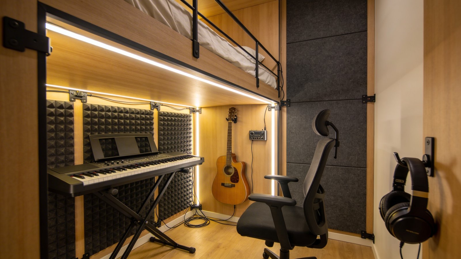 A photorealistic interior shot of a cozy music studio setup beneath a raised loft bed, featuring a keyboard synthesizer on a stand, an acoustic guitar mounted on the wall, and a small audio interface with foam acoustic panels attached to the underside of the bed frame and surrounding walls, warm ambient LED strip lighting illuminating the compact practice space, neat cable management clips running along the edges, a comfortable studio chair, and headphones hanging from a hook, captured from a side angle showing both the organized equipment layout and the loft bed structure above, natural wood tones mixed with dark gray acoustic foam, modern minimalist bedroom aesthetic