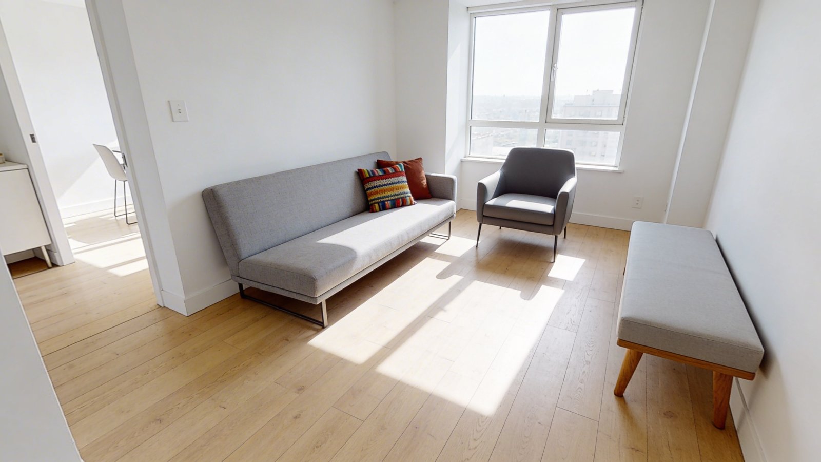 A bright, airy small apartment living room photographed from a corner angle, featuring a modern low-profile armless sofa in light gray fabric positioned against a white wall, accompanied by a sleek armless accent chair and minimalist bench with wooden legs, all arranged to showcase unobstructed sightlines across the open floor plan, with colorful throw pillows adding comfort, natural afternoon sunlight streaming through windows, pale hardwood floors, and strategic spacing between furniture pieces that emphasizes visual flow and spaciousness, shot in contemporary interior design photography style with shallow depth of field