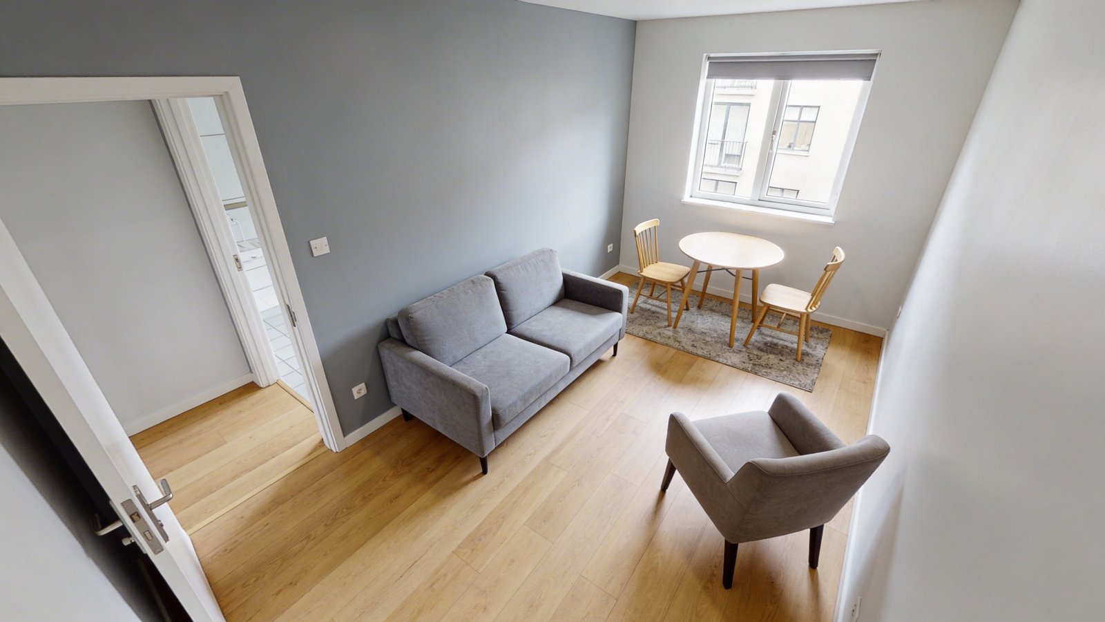 A bright modern small apartment living room photographed from a corner angle, featuring a compact two-seater loveseat in soft gray fabric perfectly proportioned to the space, a petite round dining table with two chairs in light wood against the wall, and a small-scale armchair, all arranged with ample walking space between furniture pieces, hardwood floors visible showing good flow and circulation, natural daylight streaming through a window, minimalist decor with a small area rug, demonstrating how appropriately-sized furniture creates an open and uncluttered feeling in a compact urban dwelling
