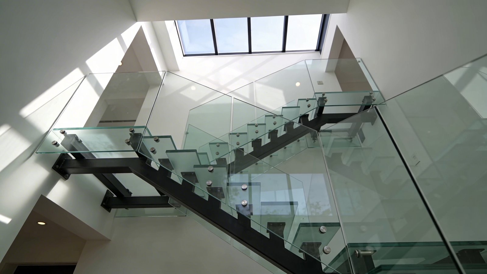 A modern attic staircase photographed from a low angle showcasing transparent tempered glass stair treads and sleek glass panel railings, with natural sunlight streaming through from a skylight above, creating beautiful light patterns and reflections on the clear glass surfaces, the staircase features minimalist black metal framework supporting the glass panels, bright and airy atmosphere with white walls, contemporary residential interior design, architectural photography, professional lighting highlighting the transparency and flow of light through multiple glass levels, shallow depth of field, high-end home renovation aesthetic
