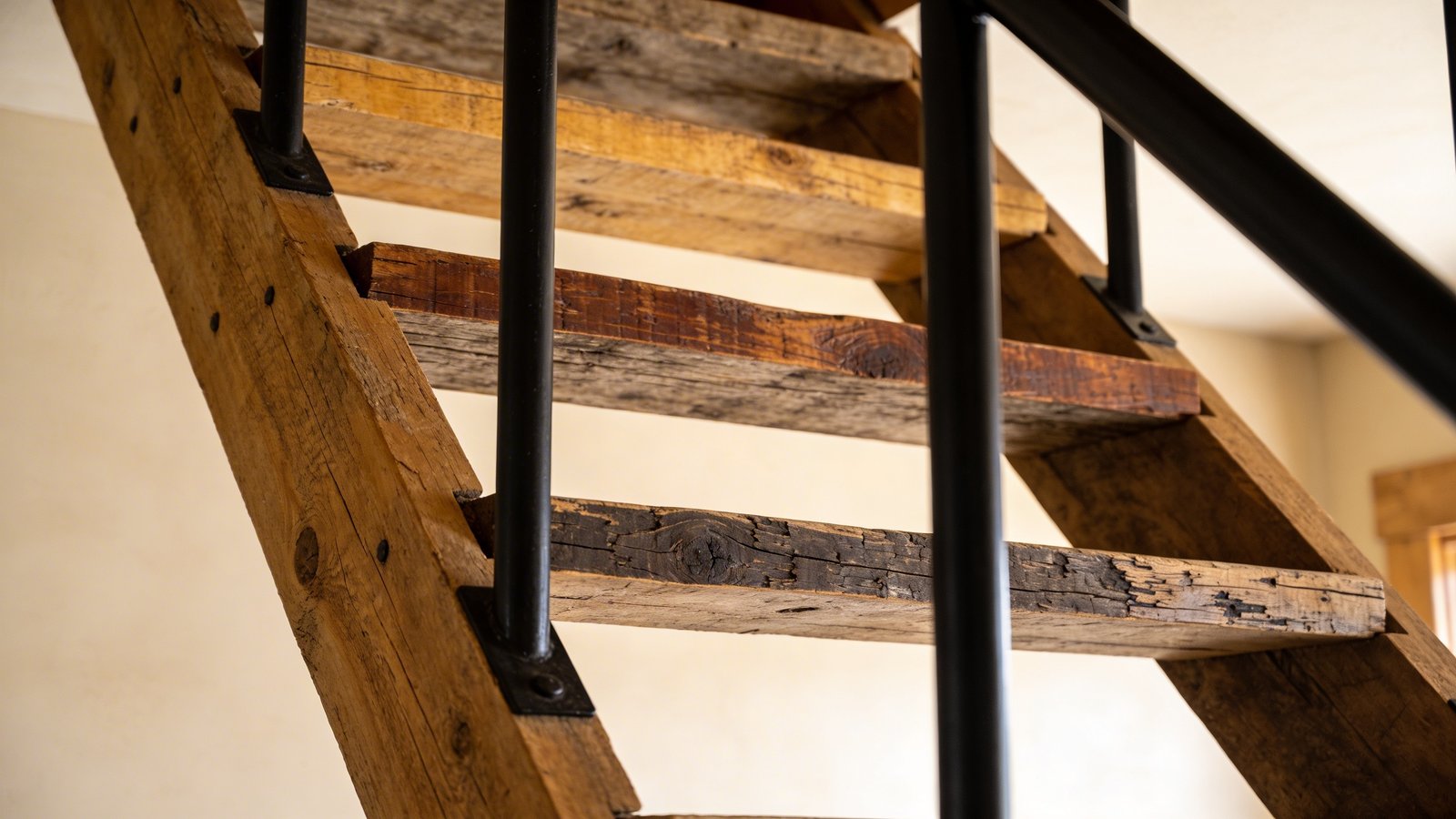 A close-up angled view of rustic reclaimed wood ladder-style attic stairs ascending into a bright space, featuring weathered barn wood treads with natural color variations from honey to deep walnut tones, visible wood grain and distressed texture, paired with matte black metal railings creating strong contrast, warm natural lighting highlighting the rough-hewn character of each wooden step, farmhouse-industrial interior design aesthetic, shallow depth of field focusing on the middle section of the stairs, warm beige walls in background