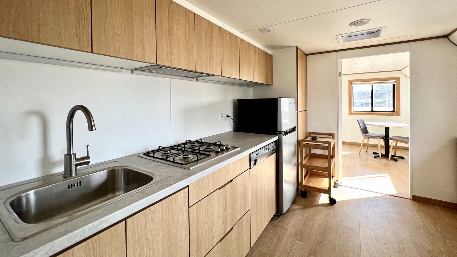 A bright, modern tiny home interior showcasing a sleek one-wall kitchen layout with all appliances and cabinets aligned along a single white wall, featuring a stainless steel sink, compact stovetop, and mini refrigerator arranged efficiently in sequence, light wood upper and lower cabinets providing ample storage, light gray countertops, a small wooden rolling cart with wheels positioned nearby for additional prep space, the opposite side of the room open and spacious with a small round dining table and chairs, natural sunlight streaming through a window, warm minimalist Scandinavian design aesthetic, shot from a three-quarter angle showing the full length of the kitchen wall and the open floor plan, professional real estate photography style