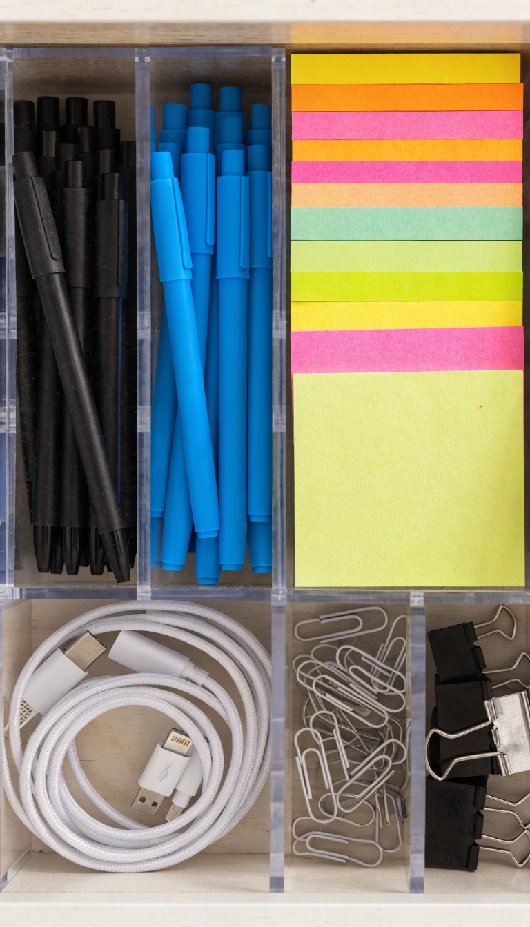 A top-down overhead photograph of an organized office desk drawer with clear acrylic dividers creating neat compartments, each section containing sorted office supplies including black and blue pens grouped together, colorful sticky notes neatly stacked, white charging cables coiled and separated, silver paperclips in one section, and small binder clips organized by size, all arranged in a modern white or light wood drawer with soft natural lighting casting subtle shadows, professional product photography style with sharp focus and clean aesthetic