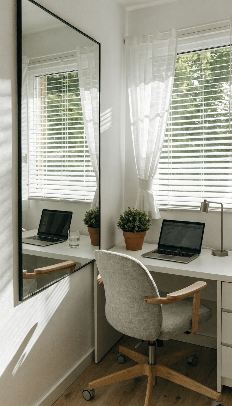 A bright, modern small corner office workspace with a large rectangular mirror mounted on the wall at seated eye level, reflecting the desk setup and creating an illusion of expanded space. The corner features a compact white desk with a laptop, small potted plant, and minimalist accessories. Natural light streams through a nearby window, bouncing off the mirror's surface and illuminating the cozy workspace. The mirror has a thin black metal frame and clearly shows the reflection of the opposite wall and desk items, demonstrating the space-doubling effect. Clean, Scandinavian-inspired design with white walls, light wood flooring, and a comfortable ergonomic chair tucked into the corner angle.