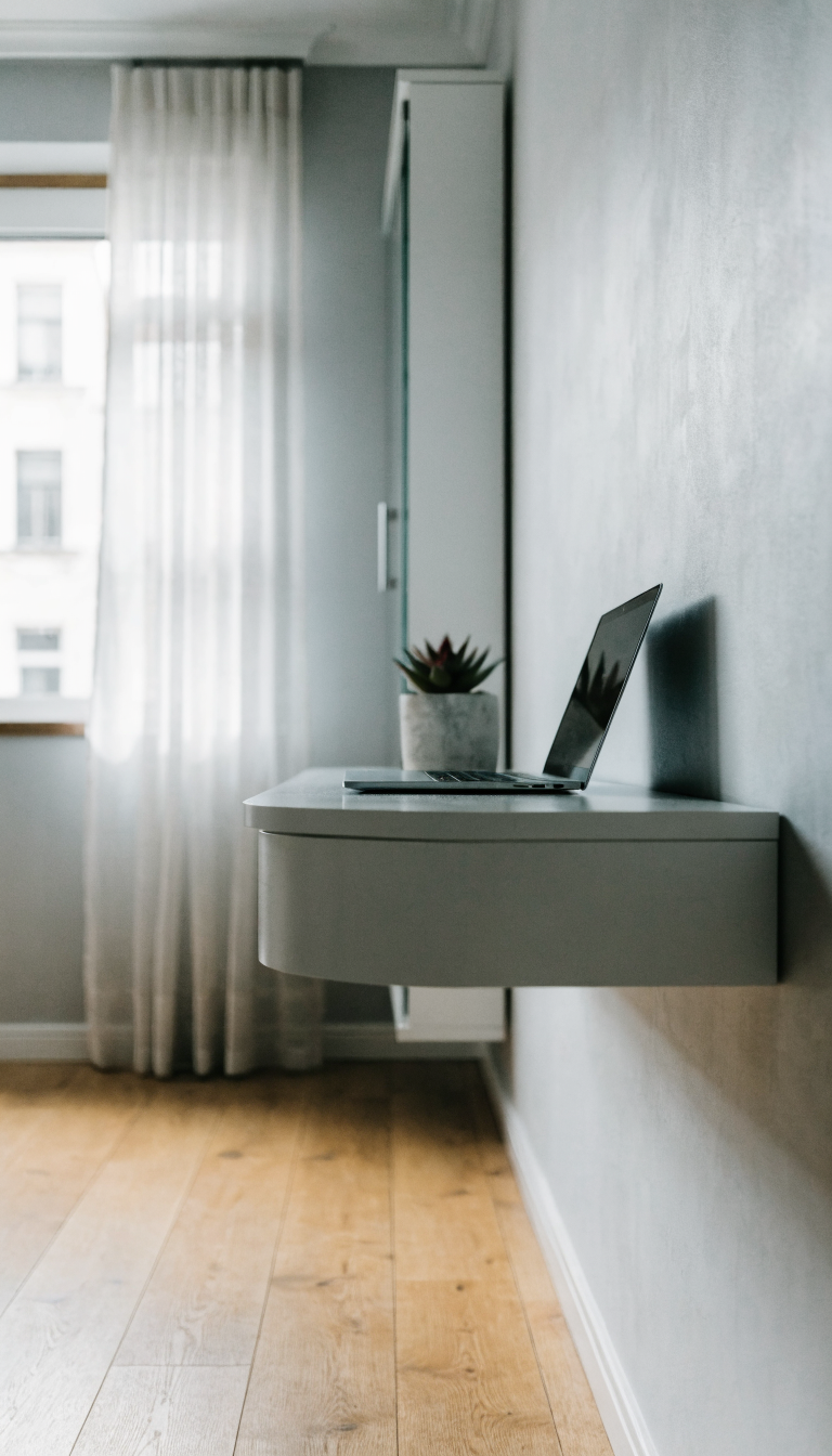 A modern minimalist white floating wall-mounted desk in a small contemporary home office, sleek and compact design attached flush against a light gray wall, clean workspace with a laptop and small succulent plant, natural daylight streaming through a nearby window, wooden floor visible below showing the space-saving benefits, Scandinavian aesthetic with neutral tones, professional interior photography style, shallow depth of field, bright and airy atmosphere