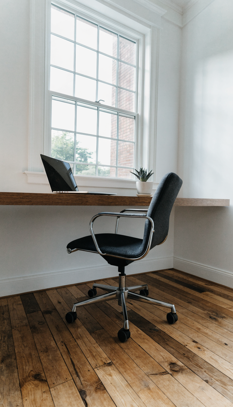 A modern minimalist home office in a small room featuring a sleek wall-mounted floating wooden desk positioned beneath a bright window with natural light streaming in, the desk has no legs and appears to float against a white wall, a comfortable office chair is tucked underneath, a laptop and small potted plant sit on the desk surface, the floor beneath is completely clear showing reclaimed hardwood flooring, the composition emphasizes the open floor space and airy feel of the room, professional interior design photography, bright and clean aesthetic, shallow depth of field